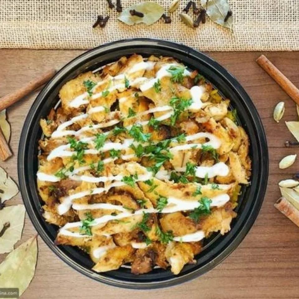 Chicken shawarma bowl with white sauce and parsley, surrounded by spices on a wooden table.