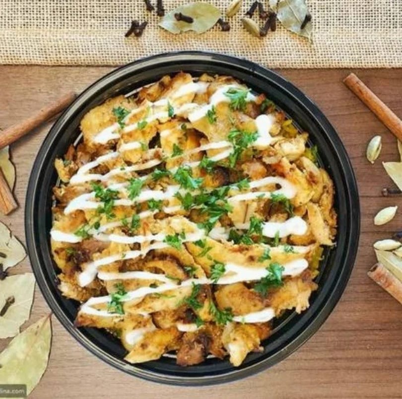 Chicken shawarma bowl with white sauce and parsley, surrounded by spices on a wooden table.