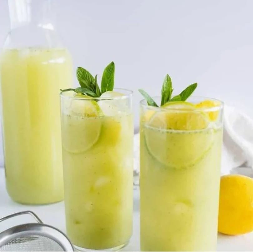 Two glasses of mint limeade with a pitcher, lemon, and strainer on a white surface.