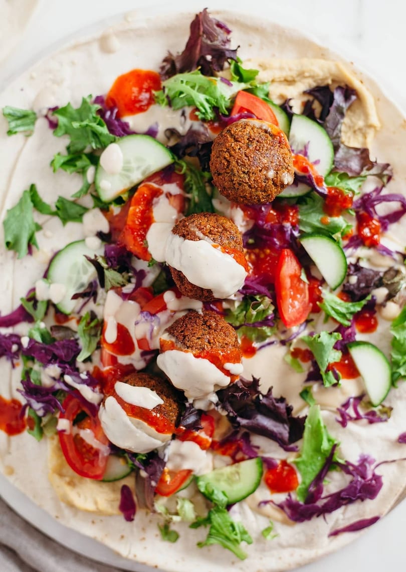 Overhead view of a falafel wrap with fresh vegetables, hummus, and sauces on a tortilla.